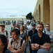 A large group of civilians and military personnel are gathered outdoors near a tan/beige building with arched architectural details, likely on a military base. The crowd appears to be attentively watching or listening to something off-camera, with many individuals standing with arms crossed. The setting and mix of uniformed and civilian attendees suggests an official event, demonstration, or briefing.