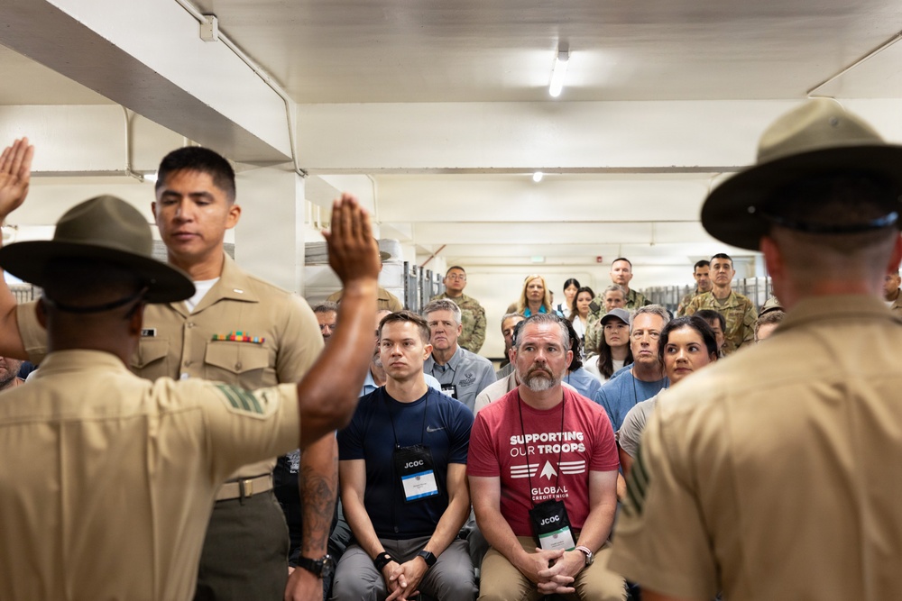 A military briefing or demonstration is taking place in what appears to be a barracks or training facility. Two drill instructors in campaign covers (campaign hats) are addressing a mixed audience of military personnel and civilians, one with a raised hand. Civilians wearing JCOC (Joint Civilian Orientation Conference) badges are seated among the audience, suggesting this is an official military orientation program.