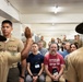 A military briefing or demonstration is taking place in what appears to be a barracks or training facility. Two drill instructors in campaign covers (campaign hats) are addressing a mixed audience of military personnel and civilians, one with a raised hand. Civilians wearing JCOC (Joint Civilian Orientation Conference) badges are seated among the audience, suggesting this is an official military orientation program.