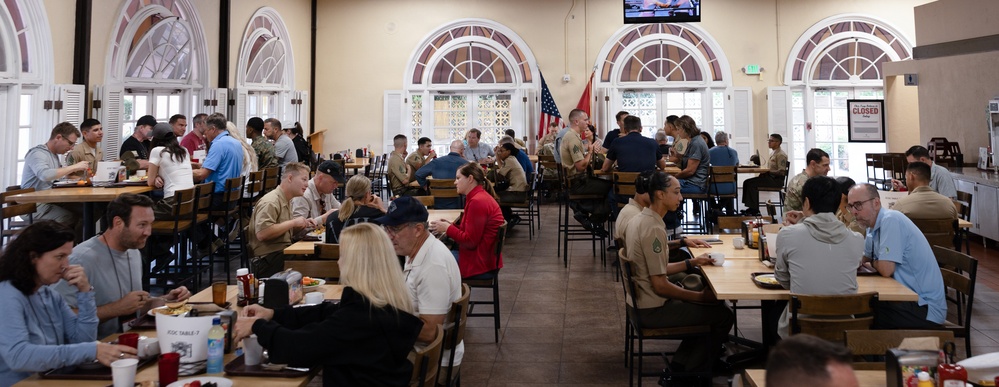 A large military dining facility (mess hall or chow hall) is filled with personnel eating meals together. The room features distinctive arched windows with white shutters and high ceilings, suggesting a formal or historic building. A mix of uniformed military personnel and civilians are seated at wooden tables throughout the space.