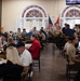 A large military dining facility (mess hall or chow hall) is filled with personnel eating meals together. The room features distinctive arched windows with white shutters and high ceilings, suggesting a formal or historic building. A mix of uniformed military personnel and civilians are seated at wooden tables throughout the space.