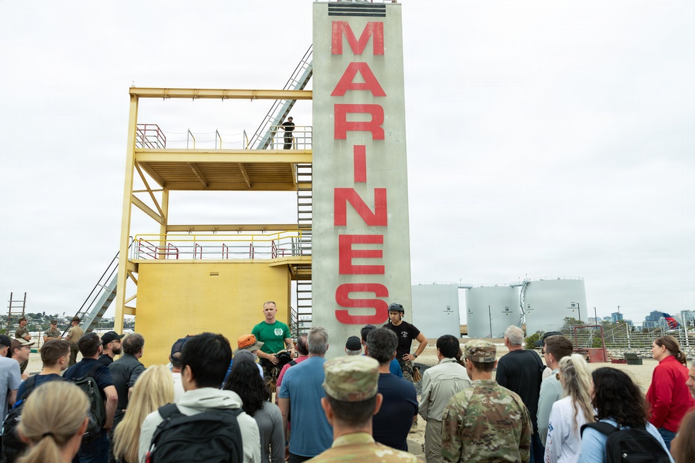 A group of civilians and military personnel are gathered at what appears to be a U.S. Marines training or recruitment facility, likely at a base near a coastal urban area. A prominent concrete pillar with 'MARINES' written in large red letters dominates the scene, alongside a yellow multi-level training tower structure. An instructor in a green shirt appears to be briefing or demonstrating to the assembled crowd.