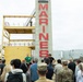 A group of civilians and military personnel are gathered at what appears to be a U.S. Marines training or recruitment facility, likely at a base near a coastal urban area. A prominent concrete pillar with 'MARINES' written in large red letters dominates the scene, alongside a yellow multi-level training tower structure. An instructor in a green shirt appears to be briefing or demonstrating to the assembled crowd.