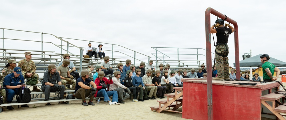 A military instructor or demonstrator stands on an elevated red platform, appearing to demonstrate equipment or techniques to a large group of civilians and military personnel seated in bleachers. The audience, many wearing military-style helmets, appears to be participating in some form of orientation, training day, or media/civilian familiarization event at an outdoor military facility. The setting appears to be a sandy training area, likely at a U.S. military base.