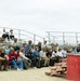 A military instructor or demonstrator stands on an elevated red platform, appearing to demonstrate equipment or techniques to a large group of civilians and military personnel seated in bleachers. The audience, many wearing military-style helmets, appears to be participating in some form of orientation, training day, or media/civilian familiarization event at an outdoor military facility. The setting appears to be a sandy training area, likely at a U.S. military base.