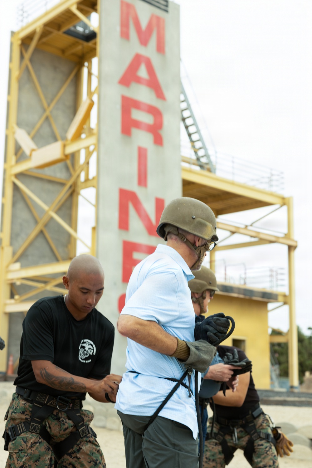 A U.S. Marine in digital camouflage uniform assists a civilian wearing a helmet and gloves with equipment at what appears to be a Marine Corps training facility. A large concrete tower with 'MARINES' written in red letters is visible in the background, along with yellow scaffolding structures. The scene suggests a civilian orientation or media event where non-military personnel are being prepared for a training exercise.