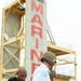 A U.S. Marine in digital camouflage uniform assists a civilian wearing a helmet and gloves with equipment at what appears to be a Marine Corps training facility. A large concrete tower with 'MARINES' written in red letters is visible in the background, along with yellow scaffolding structures. The scene suggests a civilian orientation or media event where non-military personnel are being prepared for a training exercise.