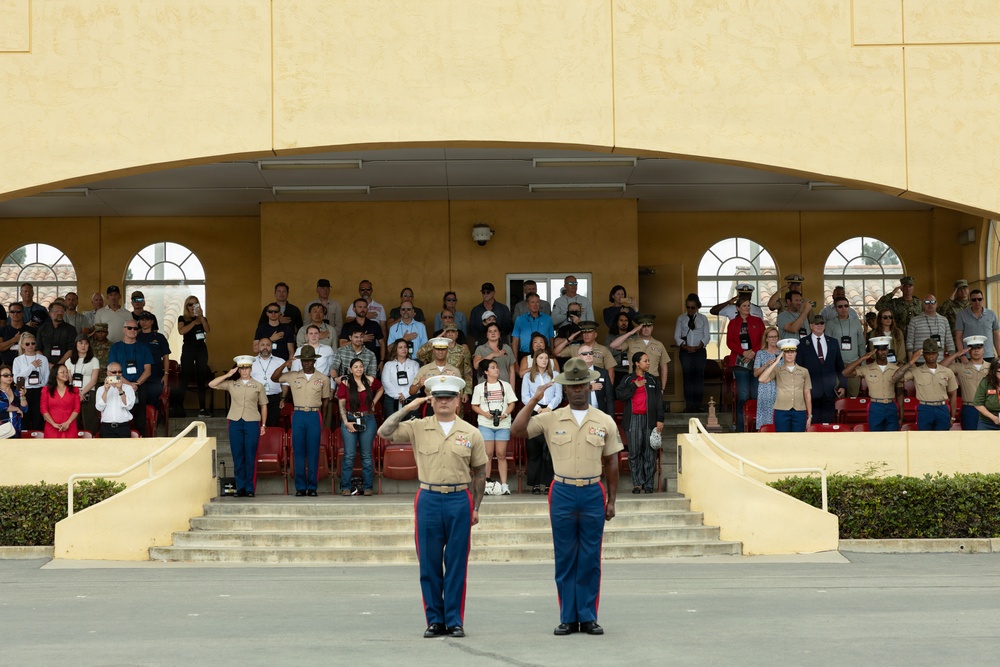A formal U.S. Marine Corps military ceremony is taking place in front of a yellow stucco building. Two Marines in dress uniforms with blue trousers and khaki blouses stand at the forefront rendering salutes, while a large crowd of military personnel and civilians in the stands behind them also salute or place hands over hearts. The setting appears to be a parade deck or ceremonial grounds at a Marine Corps installation.