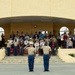 A formal U.S. Marine Corps military ceremony is taking place in front of a yellow stucco building. Two Marines in dress uniforms with blue trousers and khaki blouses stand at the forefront rendering salutes, while a large crowd of military personnel and civilians in the stands behind them also salute or place hands over hearts. The setting appears to be a parade deck or ceremonial grounds at a Marine Corps installation.