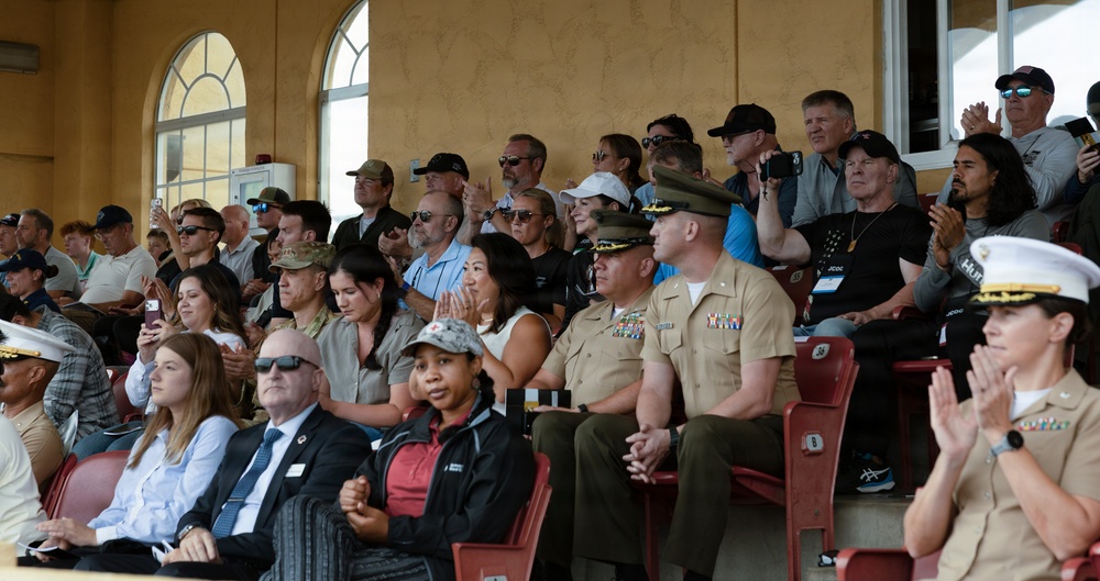 A crowd of spectators is seated in stadium bleachers, including several military personnel in uniform alongside civilians. The audience appears to be watching an event, with many people clapping and some recording with phones. The setting appears to be an indoor or covered stadium venue with a yellow stucco building visible in the background.