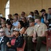 A crowd of spectators is seated in stadium bleachers, including several military personnel in uniform alongside civilians. The audience appears to be watching an event, with many people clapping and some recording with phones. The setting appears to be an indoor or covered stadium venue with a yellow stucco building visible in the background.