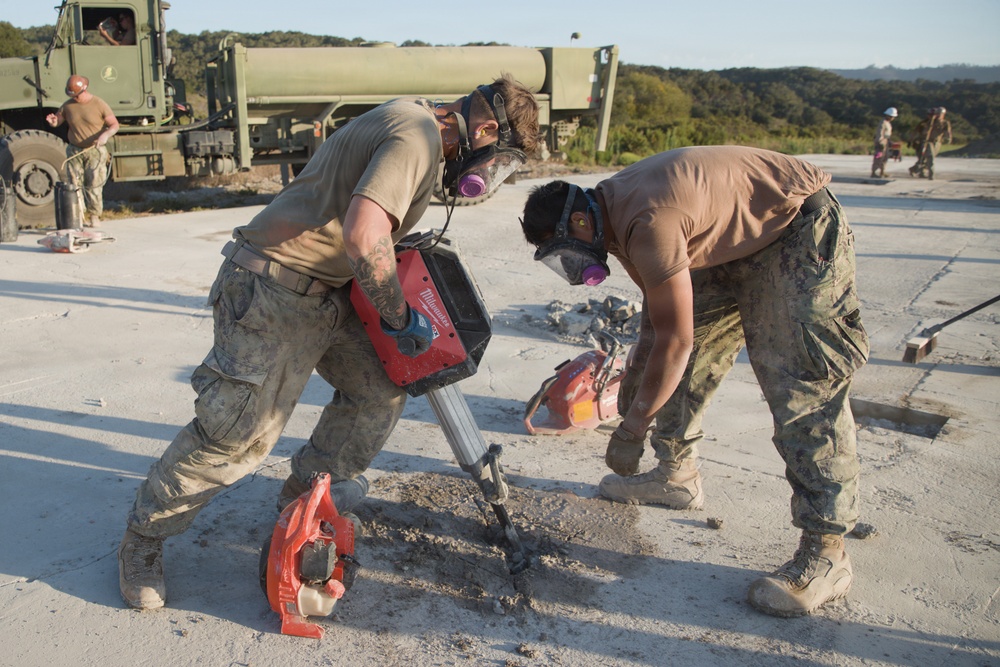 Two military personnel in camouflage uniforms and respirator masks are using a Milwaukee rotary hammer drill to break up concrete on what appears to be an airfield or runway surface. Additional personnel and a military truck are visible in the background. The scene depicts a construction or demolition operation in a field environment.