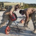 Two military personnel in camouflage uniforms and respirator masks are using a Milwaukee rotary hammer drill to break up concrete on what appears to be an airfield or runway surface. Additional personnel and a military truck are visible in the background. The scene depicts a construction or demolition operation in a field environment.