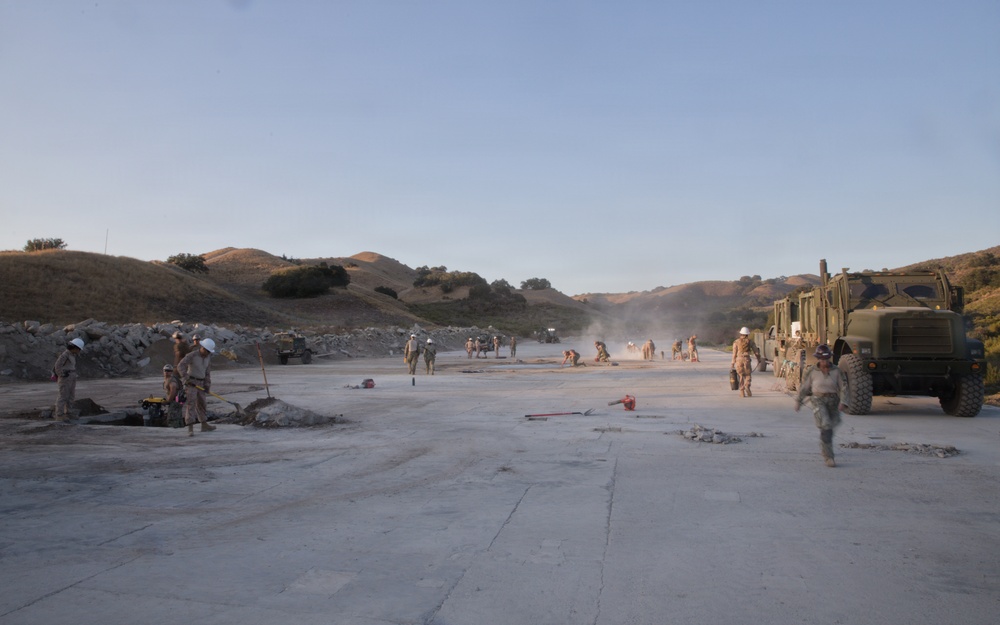 Military personnel in camouflage uniforms are engaged in construction or repair work on a large concrete surface, likely an airstrip or road, in a hilly terrain environment. A large military tactical truck (appears to be an MTVR or similar heavy vehicle) is visible on the right side. The scene suggests a combat engineering or expeditionary airfield repair exercise or operation.