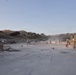 Military personnel in camouflage uniforms are engaged in construction or repair work on a large concrete surface, likely an airstrip or road, in a hilly terrain environment. A large military tactical truck (appears to be an MTVR or similar heavy vehicle) is visible on the right side. The scene suggests a combat engineering or expeditionary airfield repair exercise or operation.