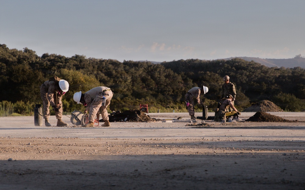 Military personnel in camouflage uniforms and white hard hats are performing construction or engineering work on a paved surface, likely an airfield or road. The workers appear to be drilling, digging, or conducting ground work using hand tools and powered equipment. The setting is an open area surrounded by dense tree line and hills in the background.