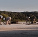 Military personnel in camouflage uniforms and white hard hats are performing construction or engineering work on a paved surface, likely an airfield or road. The workers appear to be drilling, digging, or conducting ground work using hand tools and powered equipment. The setting is an open area surrounded by dense tree line and hills in the background.