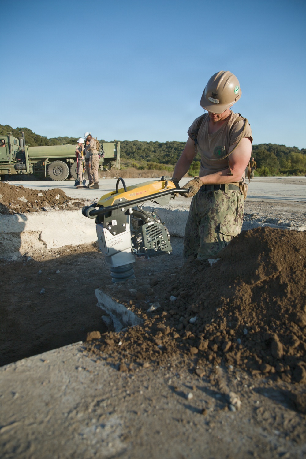 A military service member wearing camouflage uniform and a tan hard helmet operates a yellow Wacker Neuson jumping jack/rammer compactor in a trench or excavated area. The work appears to be road or surface repair/construction, with broken concrete and loose soil visible. Additional personnel and military vehicles are visible in the background.