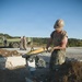 A military service member wearing camouflage uniform and a tan hard helmet operates a yellow Wacker Neuson jumping jack/rammer compactor in a trench or excavated area. The work appears to be road or surface repair/construction, with broken concrete and loose soil visible. Additional personnel and military vehicles are visible in the background.
