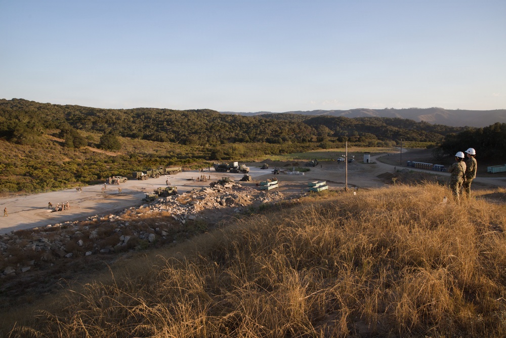 A wide-angle view of a military training or construction site in a hilly, semi-arid landscape, likely at a military installation. Two personnel wearing white hard hats observe the activity below from an elevated vantage point on a dry grass-covered ridge. Multiple military vehicles, equipment, and personnel are visible on a concrete pad area in the valley below.
