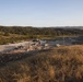 A wide-angle view of a military training or construction site in a hilly, semi-arid landscape, likely at a military installation. Two personnel wearing white hard hats observe the activity below from an elevated vantage point on a dry grass-covered ridge. Multiple military vehicles, equipment, and personnel are visible on a concrete pad area in the valley below.
