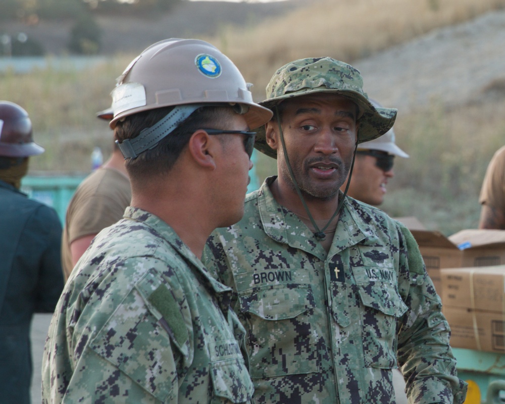 Two U.S. Navy personnel in digital camouflage uniforms (NWU Type III) are engaged in conversation at what appears to be an outdoor work or logistics site. The central figure, identified by name tape as 'BROWN,' wears a boonie hat and displays a cross insignia consistent with a Navy Chaplain. A second sailor wearing a hard hat with a green/yellow circular emblem is partially visible in the foreground.