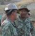 Two U.S. Navy personnel in digital camouflage uniforms (NWU Type III) are engaged in conversation at what appears to be an outdoor work or logistics site. The central figure, identified by name tape as 'BROWN,' wears a boonie hat and displays a cross insignia consistent with a Navy Chaplain. A second sailor wearing a hard hat with a green/yellow circular emblem is partially visible in the foreground.