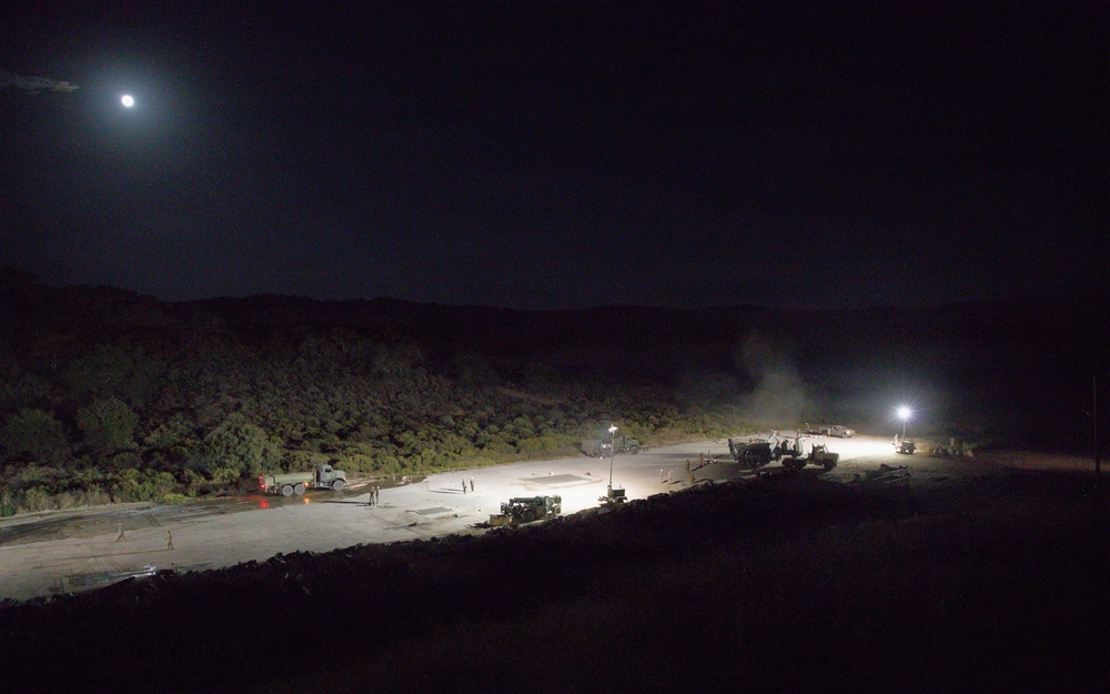 A nighttime aerial or elevated view of what appears to be a military or construction staging area, illuminated by artificial work lights and moonlight. Multiple heavy vehicles and personnel are visible on a large paved or compacted surface surrounded by dense vegetation and hills. The scene suggests active nighttime construction or engineering operations.