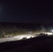 A nighttime aerial or elevated view of what appears to be a military or construction staging area, illuminated by artificial work lights and moonlight. Multiple heavy vehicles and personnel are visible on a large paved or compacted surface surrounded by dense vegetation and hills. The scene suggests active nighttime construction or engineering operations.