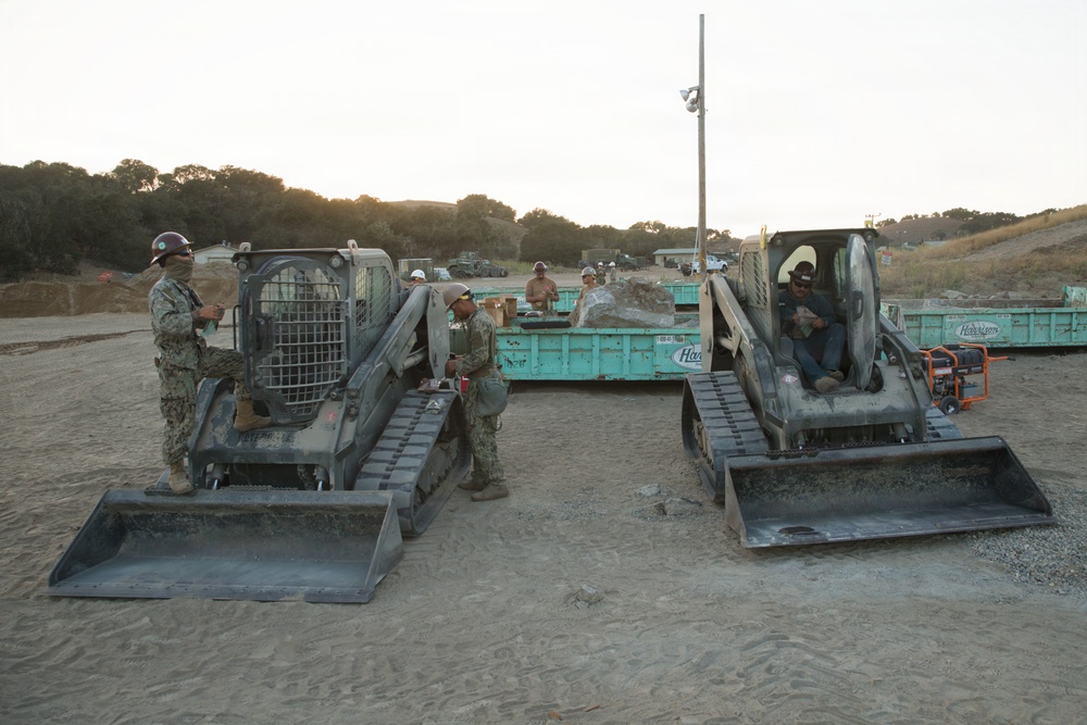 Military personnel in camouflage uniforms are operating and inspecting two tracked skid-steer loaders (likely Bobcat T-series) at an outdoor construction or training site. The scene appears to be at dusk or dawn, with rolling hills and oak trees visible in the background. Multiple personnel and additional heavy equipment, including flatbed trailers with green containers, are visible in the background.