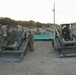 Military personnel in camouflage uniforms are operating and inspecting two tracked skid-steer loaders (likely Bobcat T-series) at an outdoor construction or training site. The scene appears to be at dusk or dawn, with rolling hills and oak trees visible in the background. Multiple personnel and additional heavy equipment, including flatbed trailers with green containers, are visible in the background.