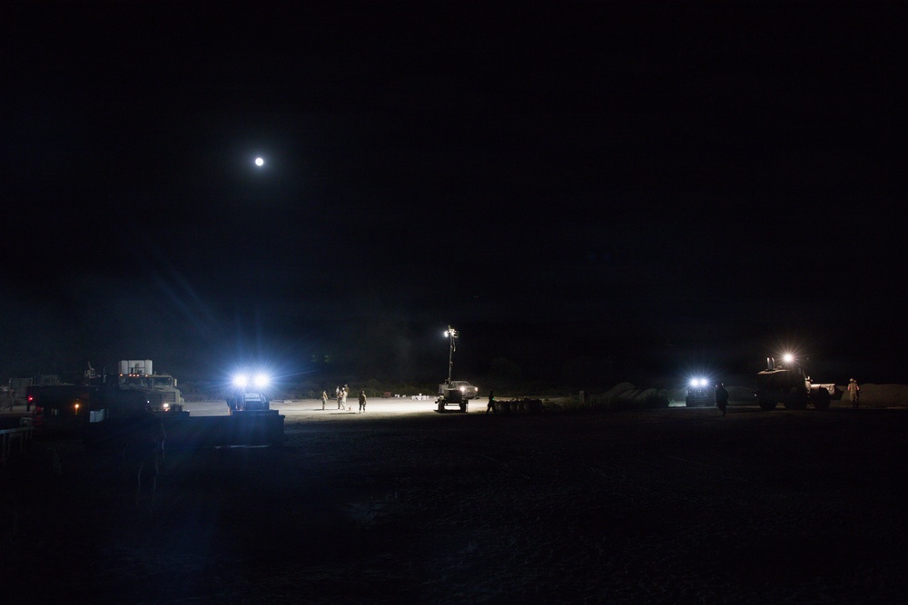A nighttime outdoor scene showing a military or construction staging area illuminated by artificial work lights and a full moon. Multiple vehicles and personnel are visible across a wide open terrain, with portable light towers providing illumination. The scene suggests an active nighttime operation or work detail in a remote or field environment.