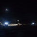 A nighttime outdoor scene showing a military or construction staging area illuminated by artificial work lights and a full moon. Multiple vehicles and personnel are visible across a wide open terrain, with portable light towers providing illumination. The scene suggests an active nighttime operation or work detail in a remote or field environment.