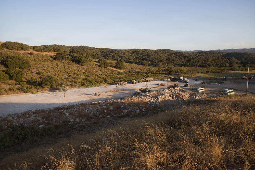 An elevated view of a military construction or engineering operation on a large concrete pad or cleared area in a hilly, semi-arid landscape. Multiple military vehicles and personnel are visible working on or around the site, with piles of rubble and debris suggesting demolition or construction activity. The setting appears to be a remote training or operational area surrounded by scrub-covered hills.