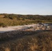 An elevated view of a military construction or engineering operation on a large concrete pad or cleared area in a hilly, semi-arid landscape. Multiple military vehicles and personnel are visible working on or around the site, with piles of rubble and debris suggesting demolition or construction activity. The setting appears to be a remote training or operational area surrounded by scrub-covered hills.