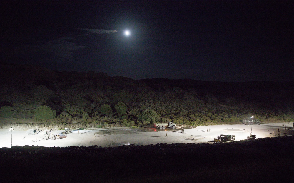 A nighttime aerial or elevated view of what appears to be a military or industrial staging area illuminated by artificial floodlights. Several vehicles, including a large truck with a flatbed trailer, are visible on a paved or compacted surface surrounded by dense vegetation and hills. A bright moon is visible in the upper portion of the dark sky.