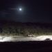 A nighttime aerial or elevated view of what appears to be a military or industrial staging area illuminated by artificial floodlights. Several vehicles, including a large truck with a flatbed trailer, are visible on a paved or compacted surface surrounded by dense vegetation and hills. A bright moon is visible in the upper portion of the dark sky.