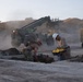 Military personnel in camouflage uniforms and white hard hats are conducting construction or repair operations on what appears to be a damaged or excavated surface, likely a runway or road. Heavy military equipment including a rough terrain forklift and a bulldozer are operating in the background amid dust clouds. The scene suggests combat engineering or airfield damage repair operations.