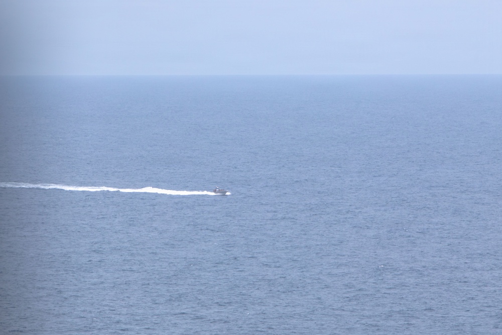 A small motorboat is traveling at speed across an open body of water, leaving a distinct white wake trail behind it. The scene is captured from an elevated vantage point, showing a vast expanse of blue-grey ocean under a hazy, overcast sky. The horizon line is clearly visible where the sea meets the sky.