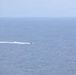A small motorboat is traveling at speed across an open body of water, leaving a distinct white wake trail behind it. The scene is captured from an elevated vantage point, showing a vast expanse of blue-grey ocean under a hazy, overcast sky. The horizon line is clearly visible where the sea meets the sky.