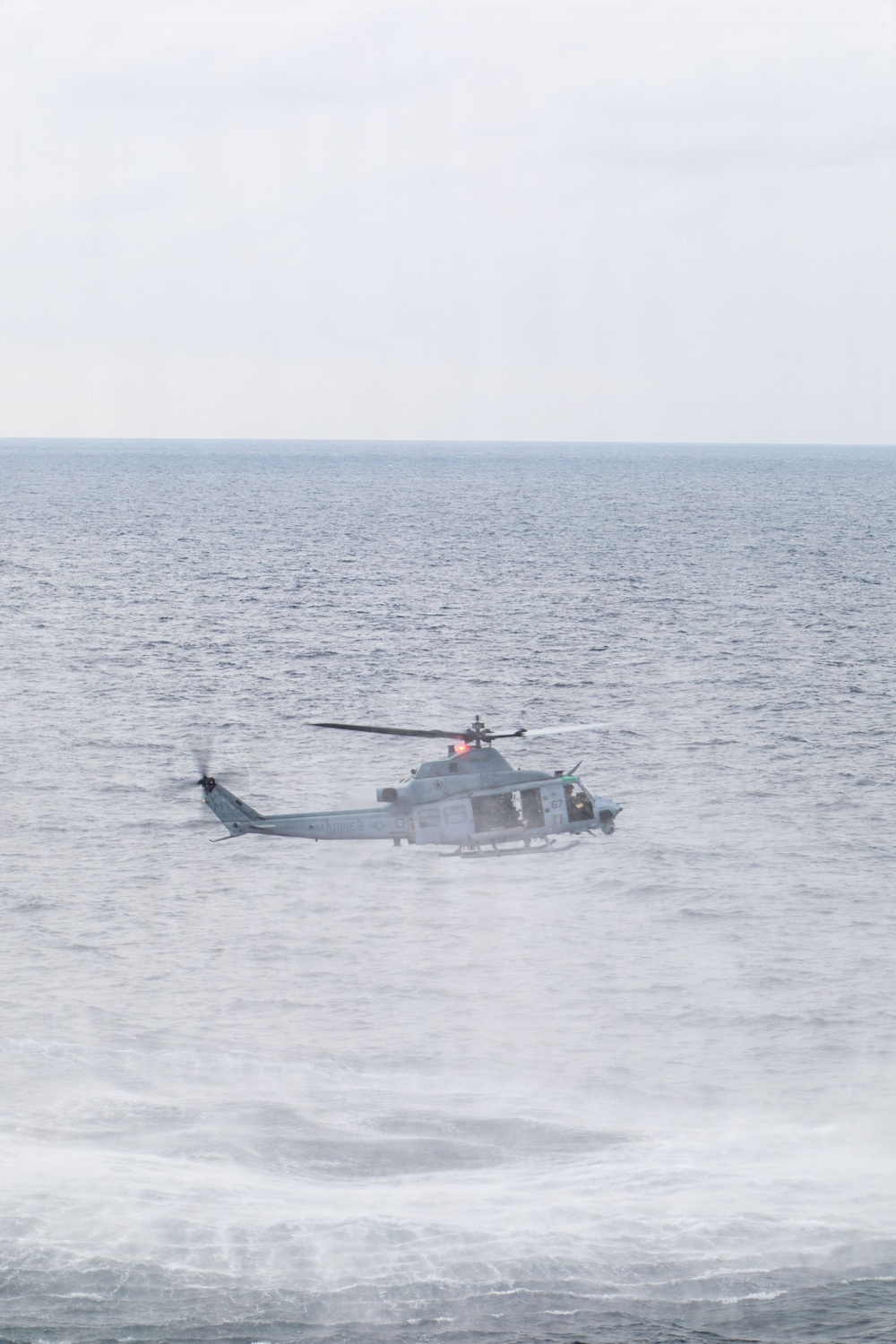 A military helicopter is flying low over open ocean waters under overcast skies. The aircraft appears to be a twin-rotor utility helicopter in a gray/light blue military paint scheme, flying at low altitude above choppy seas. The rotor wash is visibly disturbing the water surface below the aircraft.