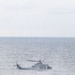 A military helicopter is flying low over open ocean waters under overcast skies. The aircraft appears to be a twin-rotor utility helicopter in a gray/light blue military paint scheme, flying at low altitude above choppy seas. The rotor wash is visibly disturbing the water surface below the aircraft.