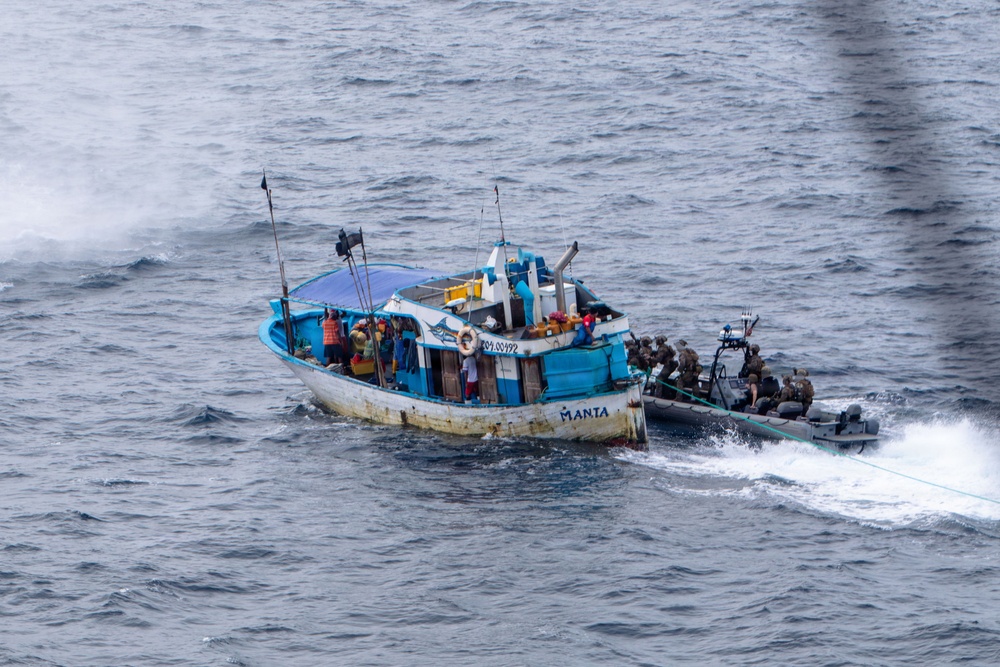 A blue and white fishing vessel named 'MANTA' is being approached and boarded by armed personnel in tactical gear arriving on a rigid inflatable boat (RIB). Multiple civilians are visible on the deck of the fishing vessel while the tactical team appears to be conducting an interdiction or law enforcement operation at sea. The image appears to be taken from an aerial or elevated vantage point, possibly from a helicopter or larger vessel.