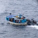 A blue and white fishing vessel named 'MANTA' is being approached and boarded by armed personnel in tactical gear arriving on a rigid inflatable boat (RIB). Multiple civilians are visible on the deck of the fishing vessel while the tactical team appears to be conducting an interdiction or law enforcement operation at sea. The image appears to be taken from an aerial or elevated vantage point, possibly from a helicopter or larger vessel.