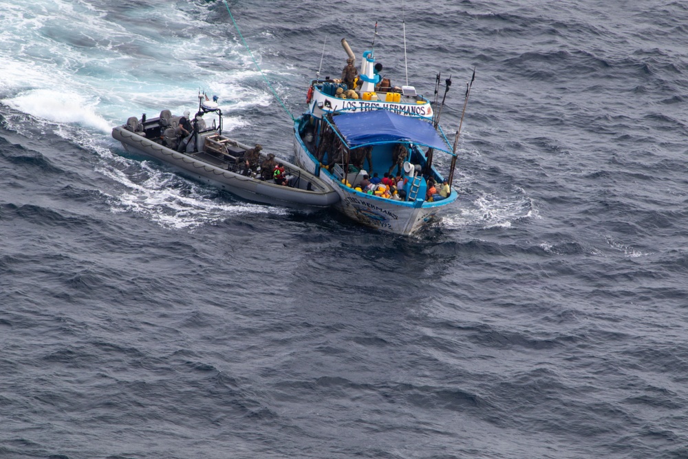 An aerial photograph shows a rigid inflatable boat (RIB) alongside a larger fishing vessel named 'Los Tres Hermanos' on open ocean waters. The RIB appears to be operated by uniformed personnel who have pulled alongside the fishing vessel, suggesting an interdiction or boarding operation. Multiple people are visible aboard the fishing vessel, which is blue and white in color.