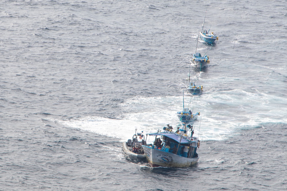 An aerial photograph shows a convoy of five blue fishing vessels traveling in a single-file line across open ocean. A smaller rigid inflatable boat (RIB) is positioned alongside the lead fishing vessel, suggesting a law enforcement or coast guard interdiction operation. The boats are leaving visible white wake trails on the gray-blue choppy water.