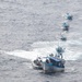 An aerial photograph shows a convoy of five blue fishing vessels traveling in a single-file line across open ocean. A smaller rigid inflatable boat (RIB) is positioned alongside the lead fishing vessel, suggesting a law enforcement or coast guard interdiction operation. The boats are leaving visible white wake trails on the gray-blue choppy water.
