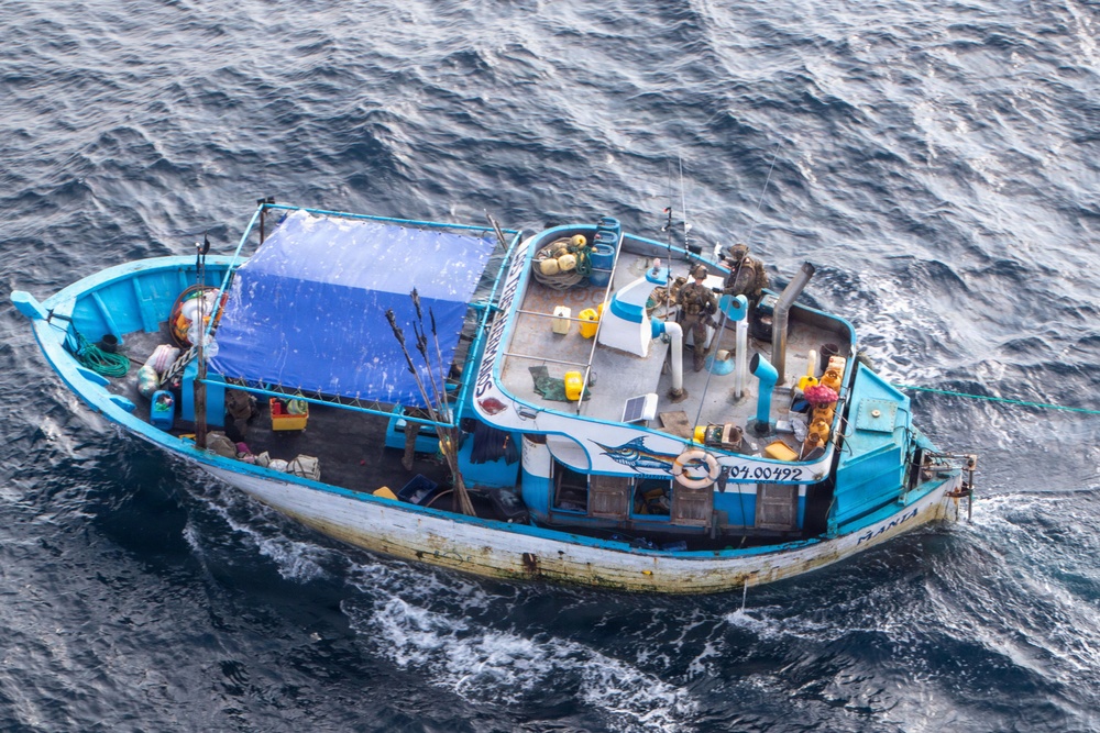 An aerial photograph of a blue and white fishing vessel at sea, identifiable by the registration number '04.00492' and a marlin logo on its hull. The boat appears to be boarded by at least two individuals in military or tactical gear on the stern deck, while other persons are visible near the bow area. A blue tarpaulin covers the mid-section of the vessel, and various fishing and deck equipment are visible throughout.