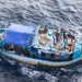 An aerial photograph of a blue and white fishing vessel at sea, identifiable by the registration number '04.00492' and a marlin logo on its hull. The boat appears to be boarded by at least two individuals in military or tactical gear on the stern deck, while other persons are visible near the bow area. A blue tarpaulin covers the mid-section of the vessel, and various fishing and deck equipment are visible throughout.
