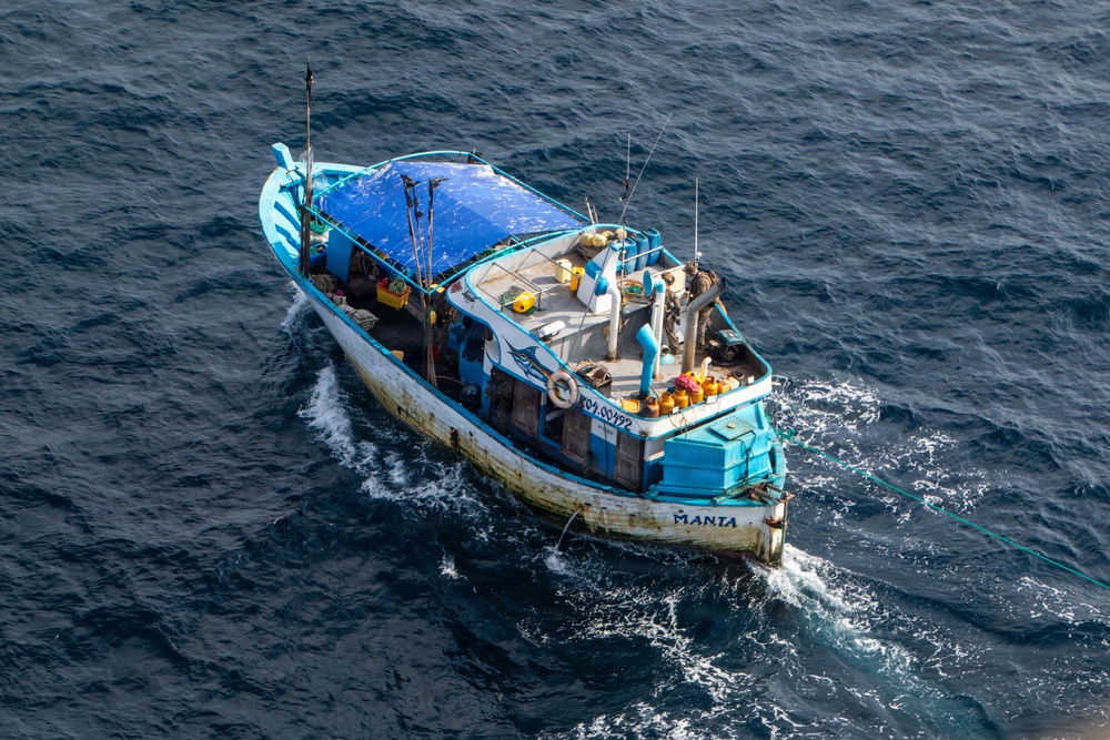 An aerial photograph of a small fishing vessel named 'MANTA' navigating open blue ocean waters. The boat displays a blue and white color scheme with visible wear and weathering on its hull. A tow line or fishing line extends from the stern into the water.